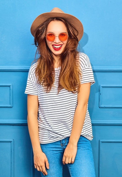 Young stylish girl model in casual summer clothes and brown hat with red lips, posing near blue wall - Fotó, kép