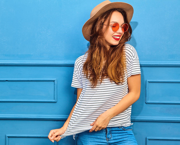 Young stylish girl model in casual summer clothes and brown hat with red lips, posing near blue wall - Foto, Bild