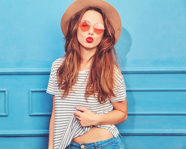 Young stylish girl model in casual summer clothes and brown hat with red lips, posing near blue wall. Giving air kiss - Φωτογραφία, εικόνα