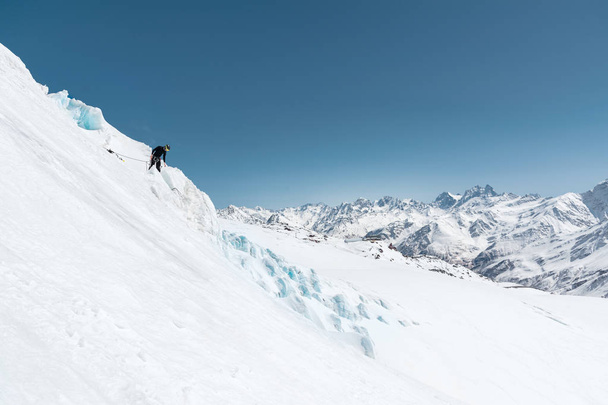 A professional mountaineer in a helmet and ski mask on insurance makes a nick-hole in the glacier against the backdrop of the Caucasian snow-capped mountains - Photo, Image