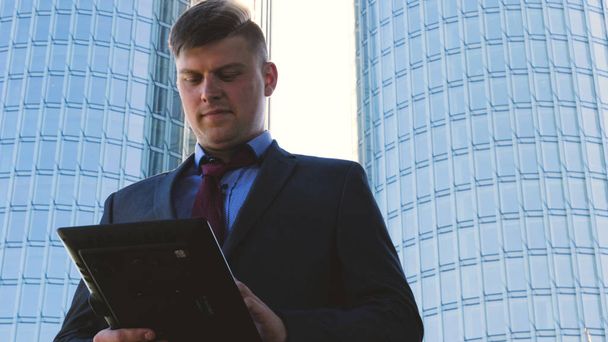 Successful handsome businessman in suit holding a tablet in his hand, working on a tablet, background of a skyscraper. Concept: communication, contacts, internet, chat, network. - Zdjęcie, obraz