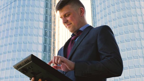 Successful handsome businessman in suit holding a tablet in his hand, working on a tablet, background of a skyscraper. Concept: communication, contacts, internet, chat, network. - Фото, зображення
