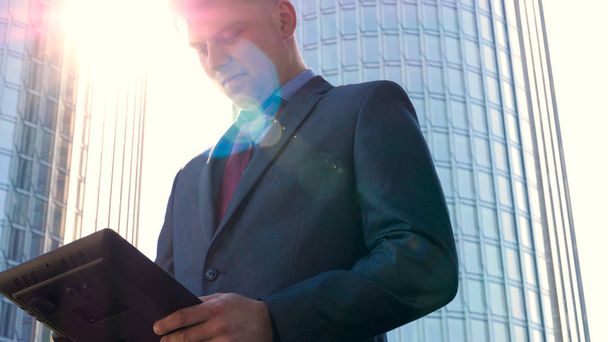 Успешный красивый бизнесмен в костюме держит планшет в руке, работает над планшетом, фоном небоскреба. Концепция: общение, контакты, интернет, чат, сеть
. - Фото, изображение