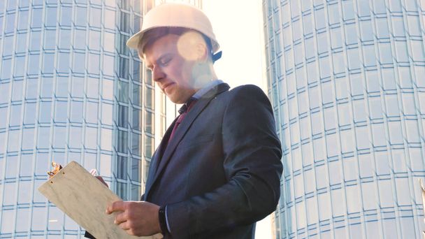 Portrait of a successful young handsome engineer, architect, builder, businessman, wearing a white helmet, in a suit, writing on a notebook, skyscraper background. - Photo, Image