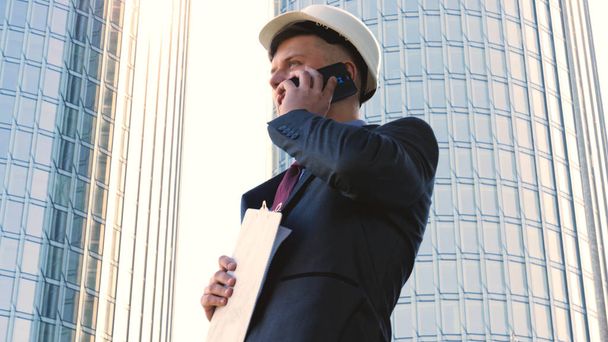 Portrait of a successful young handsome engineer, architect, builder, businessman, wearing a white helmet, in a suit, writing on a notebook, skyscraper background. - Foto, imagen