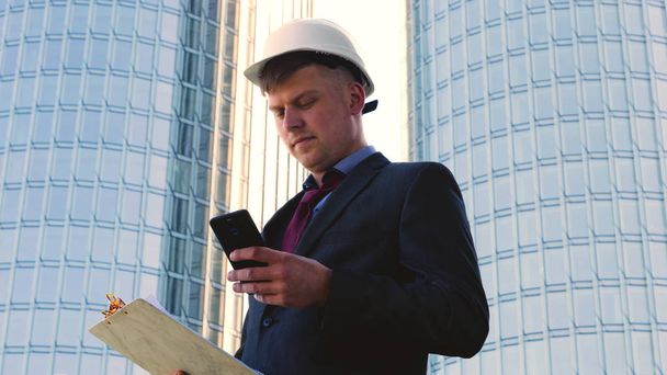 Portrait of a successful young handsome engineer, architect, builder, businessman, wearing a white helmet, in a suit, writing on a notebook, skyscraper background. - Valokuva, kuva