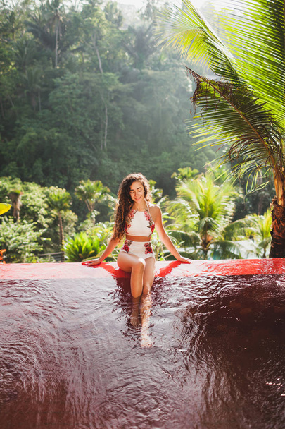 Bella ragazza abbronzata seduta sul bordo della piscina privata di lusso con splendida vista sulla giungla in mattinata di sole
 - Foto, immagini