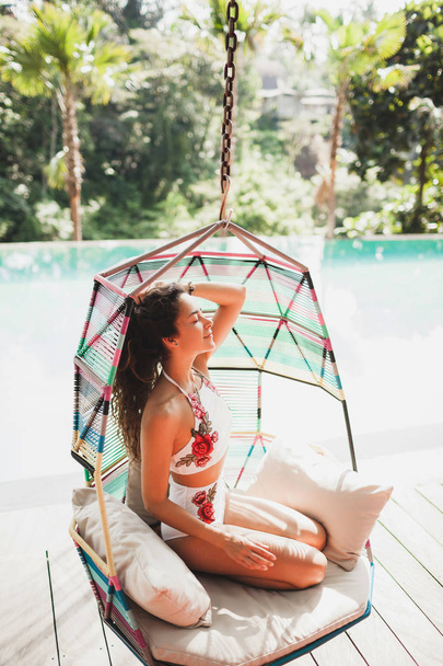Woman in white swimsuit enjoying in hanging chair swing on poolside in luxury hotel. Morning sunny mood - Foto, afbeelding