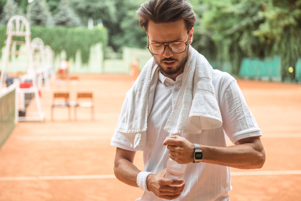 schöner Tennisspieler mit Handtuch und Flasche Wasser auf dem Tennisplatz - Foto, Bild
