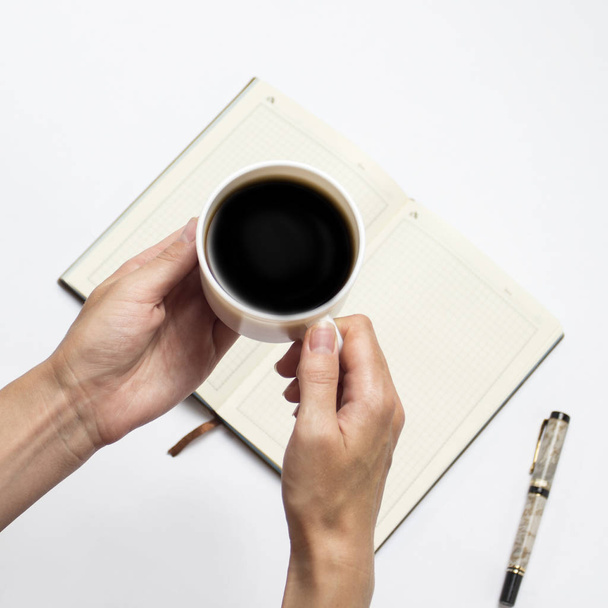 Mão feminina com uma xícara de café preto e um diário aberto com páginas limpas, uma caneta em um fundo claro. Deitado plano, vista superior
. - Foto, Imagem