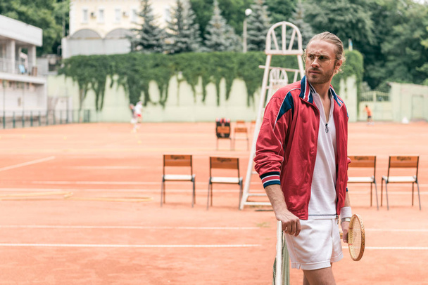 όμορφος Καυκάσιος τενίστας με ρακέτα στέκεται στο ΝΕΤ στο γήπεδο τένις - Φωτογραφία, εικόνα