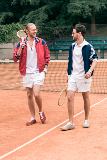 cheerful sportive friends with wooden rackets walking on tennis court - Photo, Image