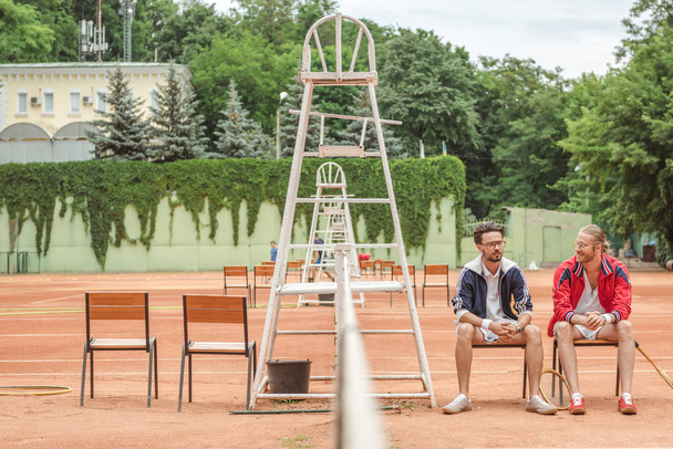 спортивні друзі з дерев'яними ракетками, що сидять на стільцях на тенісному корті
 - Фото, зображення