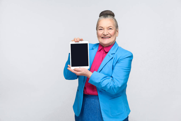 Aged woman holding and showing tablet screen with toothy smiling and satisfied face on gray background  - 写真・画像