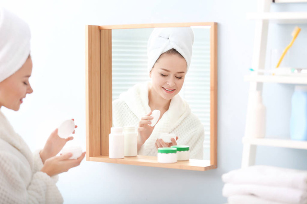 Young beautiful woman using cream in front of mirror indoors - Photo, Image