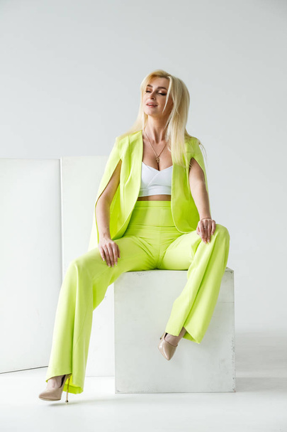 Fashion studio portrait of beauty blond model in white room interior. Shot of beauty young woman wearing green casual suit and white shirt - Zdjęcie, obraz