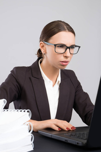 Young attentive business woman sitting at the desk with papers and books and using laptop - Foto, Bild