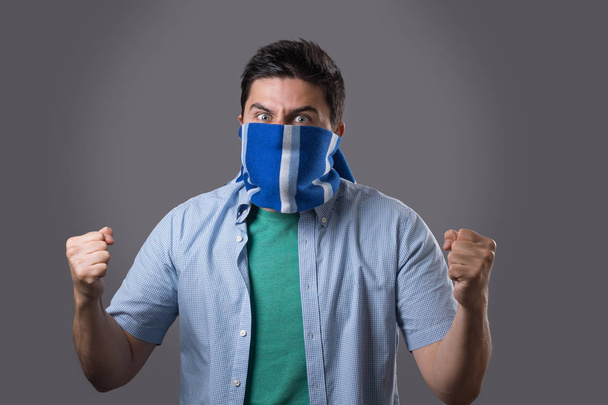 Portrait of aggressive sports fan with blue scarf covering his face isolated on grey background. Football hooligan concept - Photo, Image