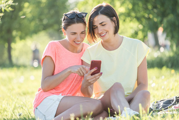 Outdoor portrait of two cheerful girls sitting on lawn, using smartphone and having good time together in park at sunny day. - Foto, Bild