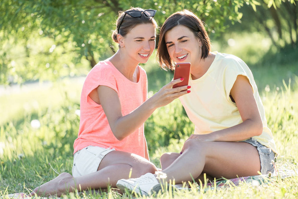 Outdoor portrait of two cheerful girls sitting on lawn, using smartphone and having good time together in park at sunny day. - Foto, Imagen