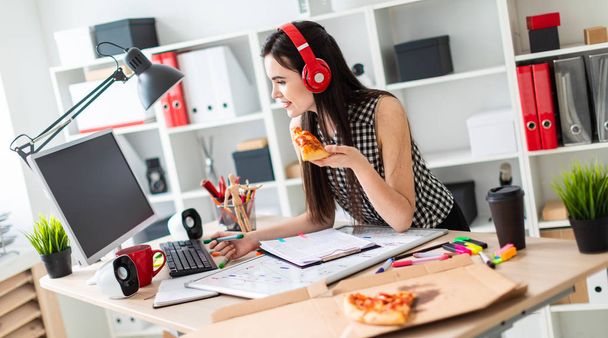 Egy fiatal lány állandó közelében egy táblázat, a zöld jelölőt és egy darab pizza tartja a kezében. Mielőtt a lány az asztalra egy mágneslemez. A lány fején a fejhallgató viselése. - Fotó, kép