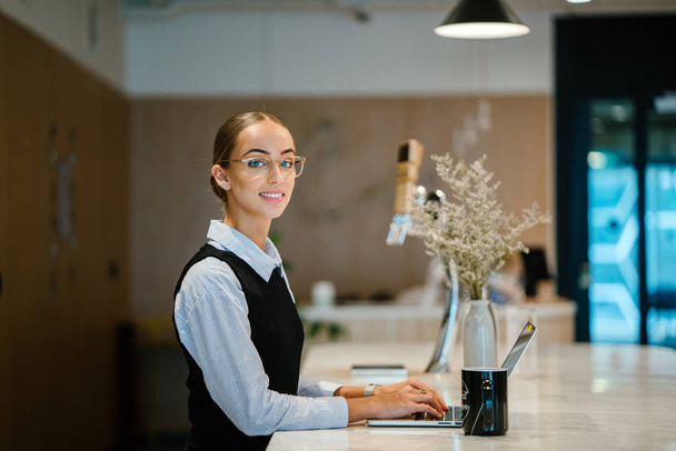 young businesswoman wearing eyeglasses smiling working on laptop sitting at table, side view - Фото, зображення