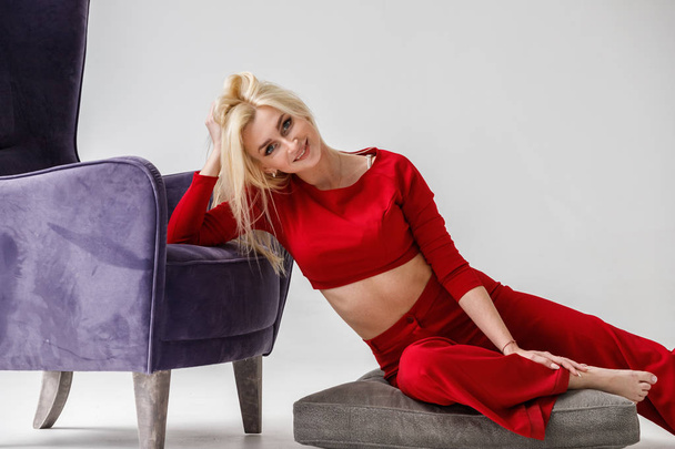Fashion studio portrait of beauty blond model in white room interior with purple armchair. Shot of beauty young woman wearing red casual suit  - Φωτογραφία, εικόνα