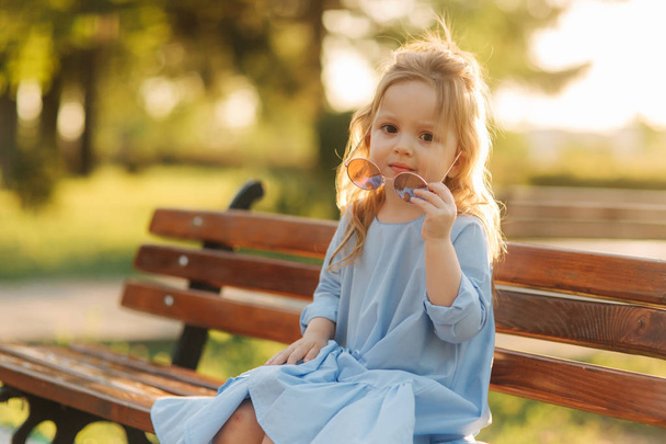 Little girl model in blue dress and sun glasses sits on a bench in the park - Photo, Image