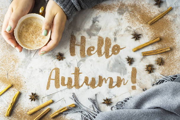 Hallo, Herbst. Der Satz ist mit gemahlenem Zimt auf grauem Hintergrund geschrieben. neben einer Tasse Cappuccino in weiblicher Hand. Draufsicht, flache Lage - Foto, Bild
