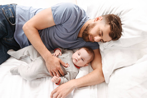 Image of handsome young man father dad sleeping in bed with his little child son indoors at home. - Foto, Imagem