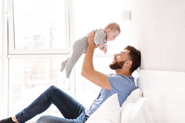 Photo of handsome happy young man father dad having fun with his little baby child son indoors at home. - Фото, зображення