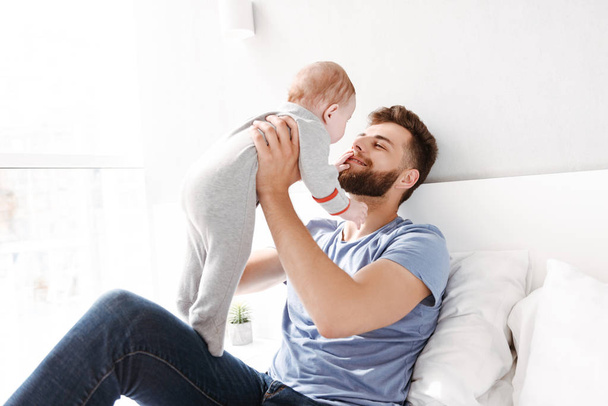 Photo of cheerful young man father dad having fun with his little baby child son indoors at home. - Foto, Bild