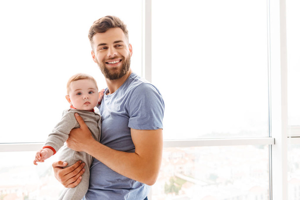 Image of handsome young cheerful bearded man holding his little son child in hands looking aside. - Φωτογραφία, εικόνα