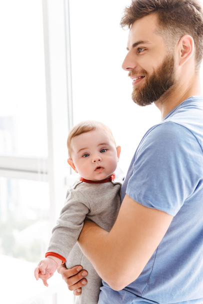 Image of handsome young cheerful bearded man holding his little son child in hands looking aside. - 写真・画像