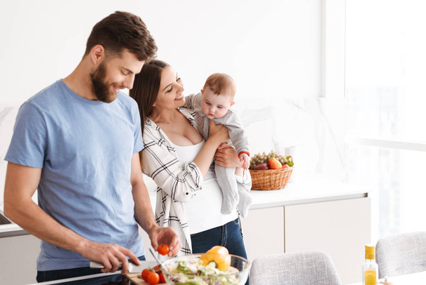 Photo of amazing young loving couple parents with their little baby son cooking in kitchen. - Photo, image