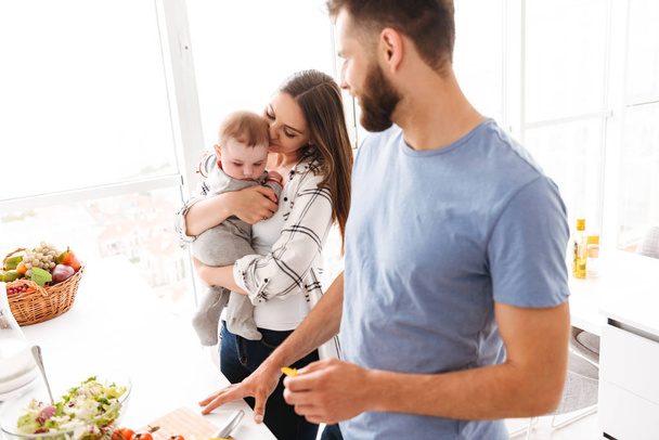 Image of happy young loving couple parents with their little child baby son cooking in kitchen. - Foto, Imagem
