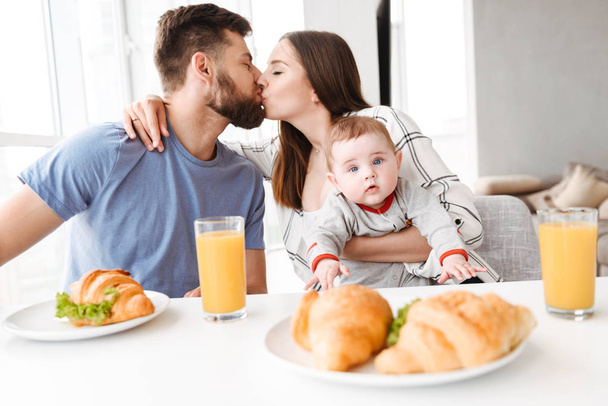 Photo of amazing young loving couple parents with their little baby son eating in kitchen kissing have a breakfast. - Foto, imagen