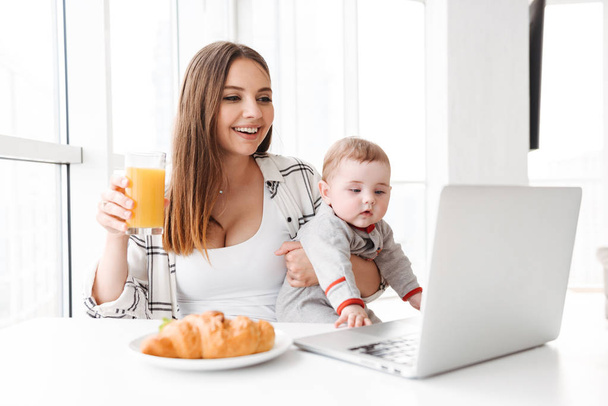 Image of happy young woman mother using laptop computer with her little child son indoors at home have a breakfast. - Fotoğraf, Görsel