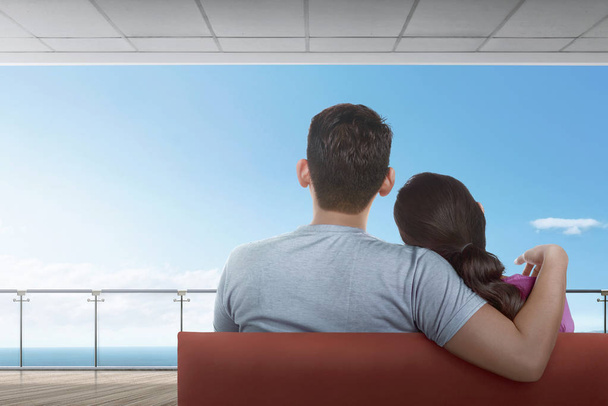 Rear view of asian couple sitting on couch and enjoying ocean view from balcony  - Foto, Imagen