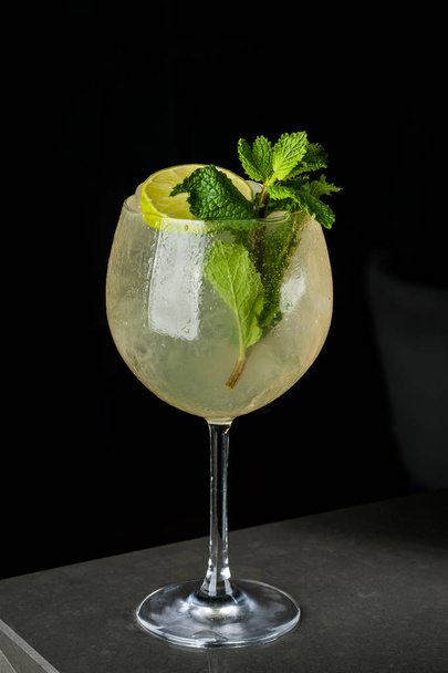 Yellow refreshing drink with lemon and mint in a glass of ice. Cocktail on a black background with bubbles and dew - 写真・画像