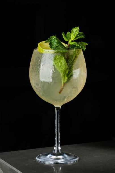 Yellow refreshing drink with lemon and mint in a glass of ice. Cocktail on a black background with bubbles and dew - Foto, Imagen
