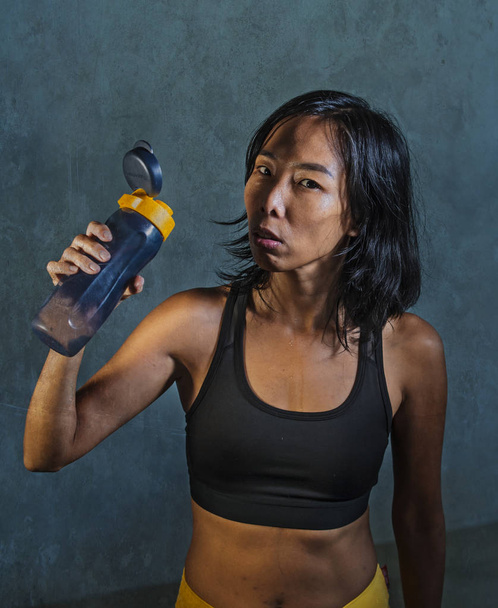 portrait of young athletic and fit Asian Korean woman in fitness top holding drinking water bottle posing cool in bad girl defiant attitude isolated on dark background in gym sport workout concept - 写真・画像