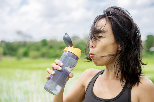young healthy sporty Asian Chinese woman drinking water bottle after fitness training and running workout outdoors on green field landscape background in sport and well being concept - Foto, immagini
