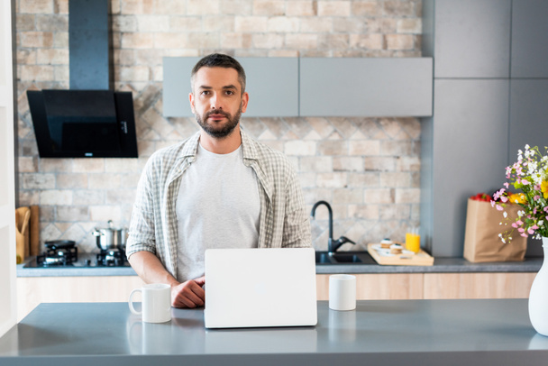 retrato de hombre barbudo de pie en el mostrador con ordenador portátil y tazas de café y mirando a la cámara en la cocina
 - Foto, Imagen