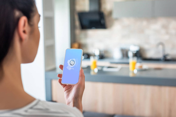 selective focus of woman holding smartphone with shazam logo on screen in kitchen - Foto, Bild