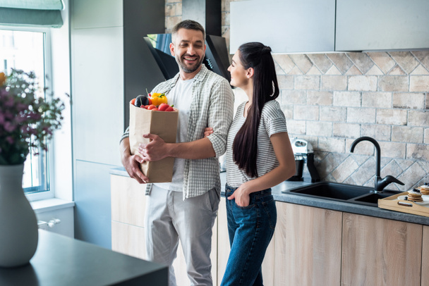 coppia allegra con pacchetto di carta pieno di verdure fresche in piedi in cucina a casa
 - Foto, immagini