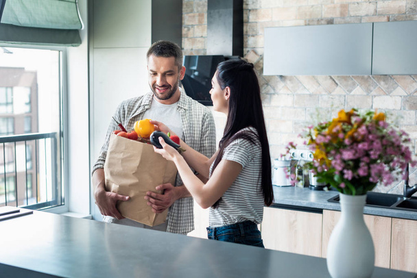 married couple with paper package full of fresh vegetables for dinner in kitchen at home - Fotoğraf, Görsel
