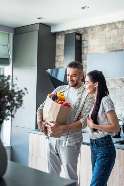 boldog házaspár papír csomag tele friss zöldségekkel, vacsora otthon a konyhában - Fotó, kép