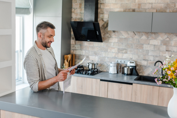 side view of smiling bearded man reading newspaper while standing at counter in kitchen - Foto, Imagen