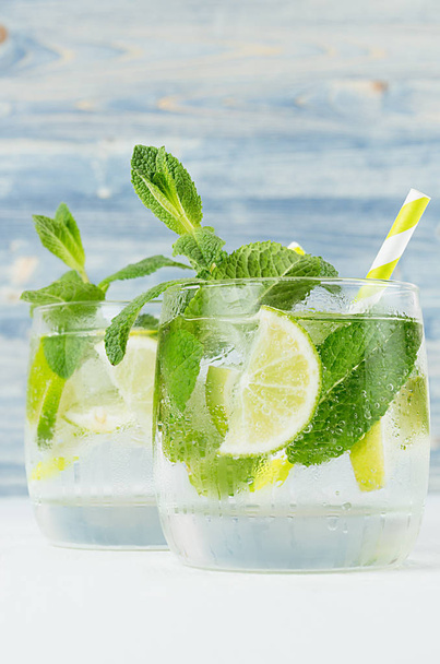 Fresh cold summer beverage gin and tonic with lime, leaf mint, straw, ice cubes, soda on light blue wooden background, closeup. - Fotó, kép
